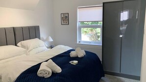 Una cama bien hecha con ropa de cama blanca y azul, una lámpara de noche, un cuadro en la pared y una ventana con vistas a la vegetación.