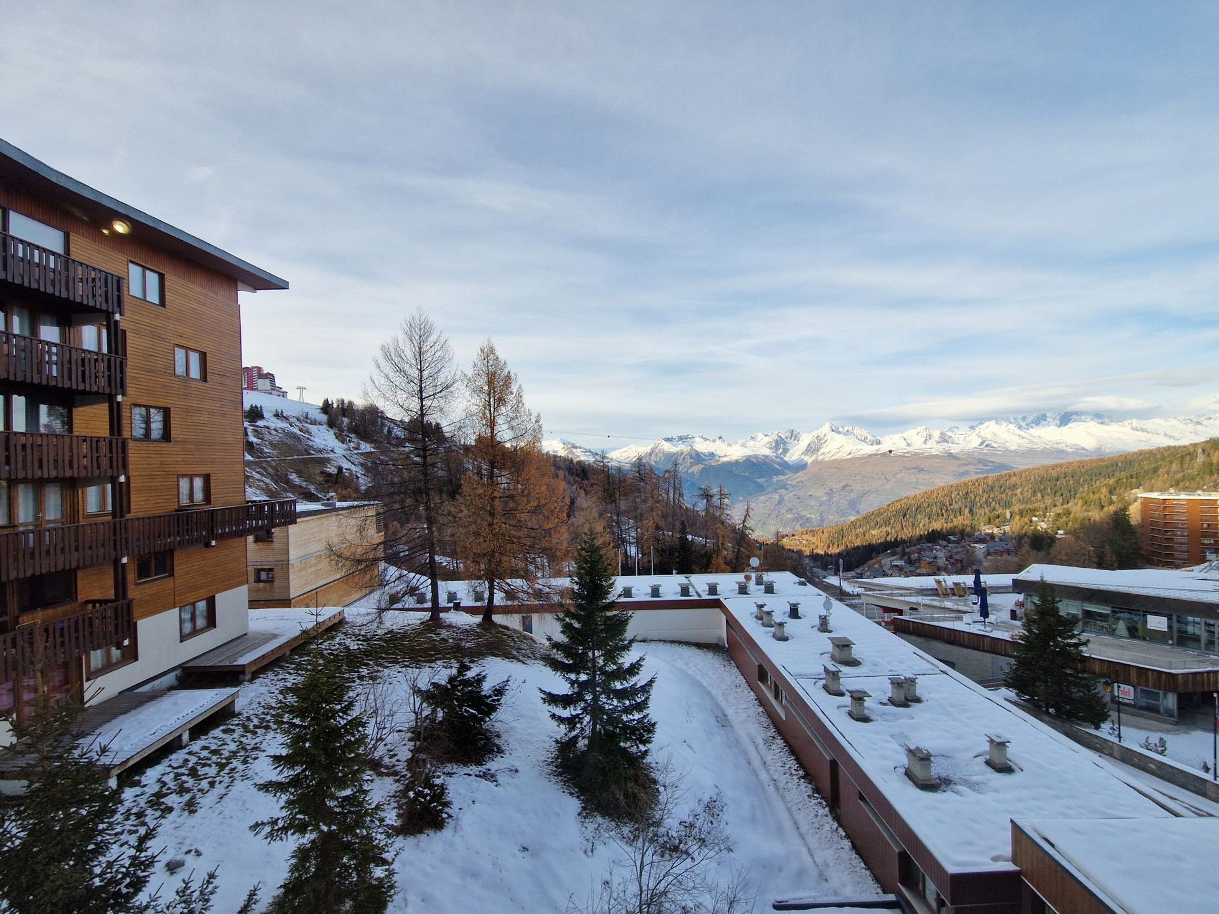 Ein modernes Gebäude mit mehreren Balkonen, einem verschneiten Innenhof und Blick auf schneebedeckte Berge im Hintergrund.
