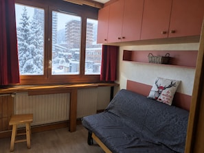A bedroom with a bed, a window with curtains, a radiator, a wooden stool, and a shelf with a basket.