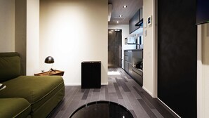 A narrow, modern hotel room with a green sofa, a small table with a lamp, a black trash bin, and a kitchen area with cabinets and a microwave. - Real Life SANGENJAYA 1BR  2Pax Shibuya (Meguro)