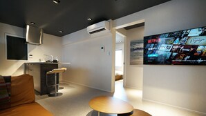 Een moderne hotelkamer met kitchenette, een grote wandtelevisie waarop verschillende programma's worden vertoond, en een gezellige zithoek met een bank en een klein houten tafeltje.