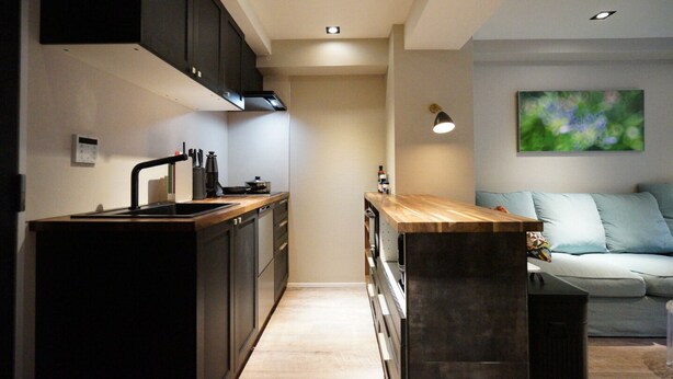 A modern kitchen with dark cabinetry, a wooden countertop, and a built-in oven. There is a small island with a sink and a light fixture above it. A sofa with a cushion is visible in the background.