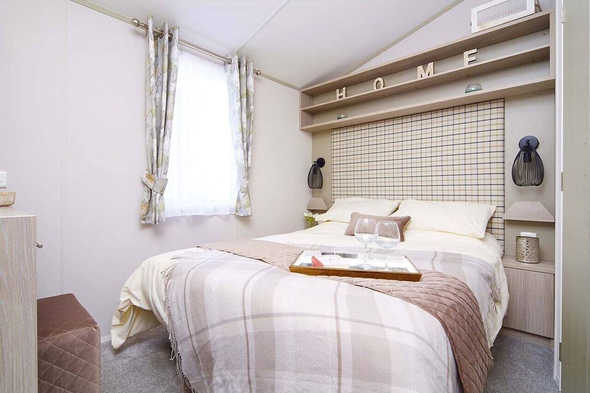 Un dormitorio con cama, mesita de noche, estante con la palabra 'HOME' escrita, ventana con cortinas y una lámpara de noche.