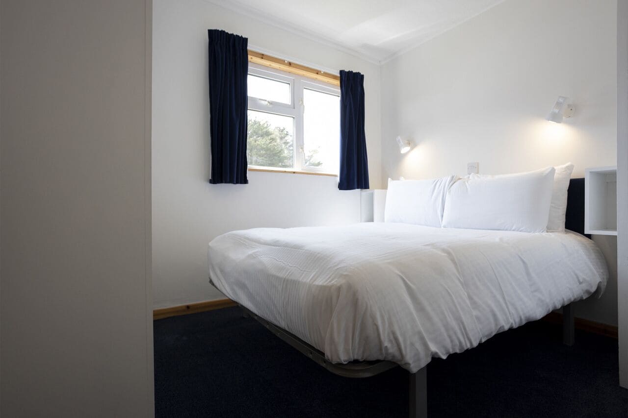 A neatly made bed with white bedding in a room with white walls, a window with dark curtains, and a bedside table with a lamp.
