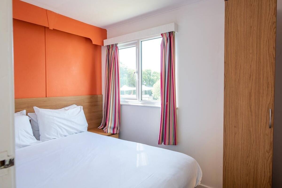 Una habitación de hotel con una cama, un ventanal con cortinas a rayas y una pared de acento naranja.