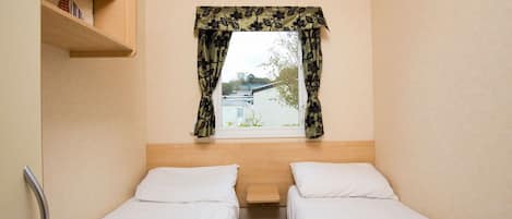 A small, single-room hotel room with two beds, a window with floral curtains, a wooden shelf with books, and a small table between the beds.