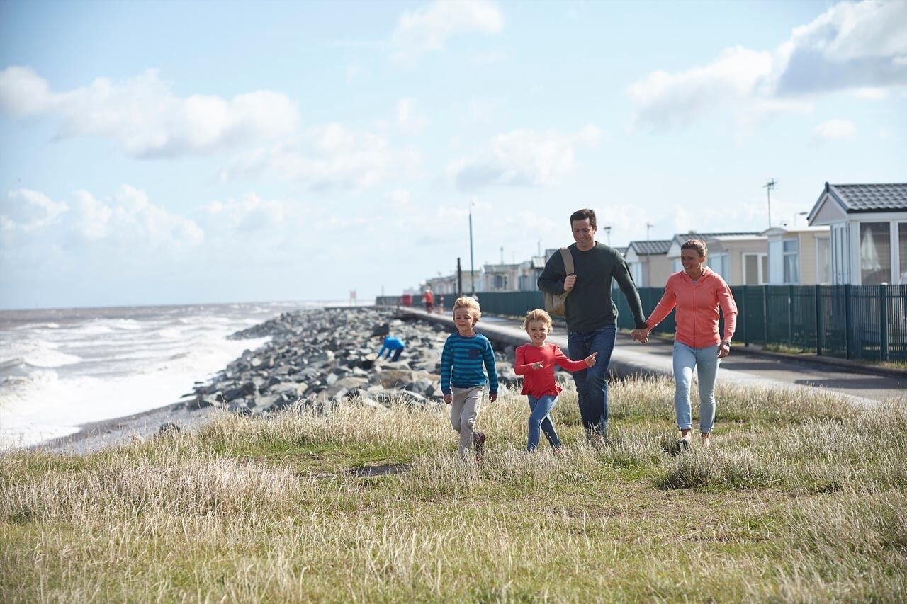 A family walking along a grassy path beside a rocky beach with houses in the background.