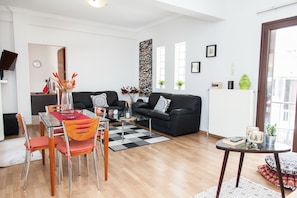 A modern living room with a black sofa, a glass coffee table, and a dining table with chairs. There are potted plants and a large abstract painting on the wall.