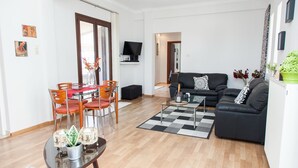 Una sala moderna con un sofá negro, una mesa de comedor con sillas, un televisor montado en la pared y una planta verde en maceta.