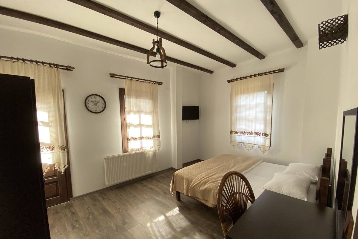 Une chambre avec un plancher en bois, un lit recouvert d’une couvre-lit beige, une chaise en bois, une commode, une horloge au mur et deux fenêtres avec des rideaux.