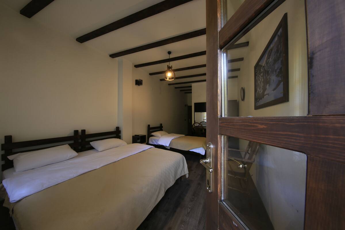 Une chambre d’hôtel avec trois lits simples, des poutres en bois au plafond, un tableau encadré au mur et une porte en bois.