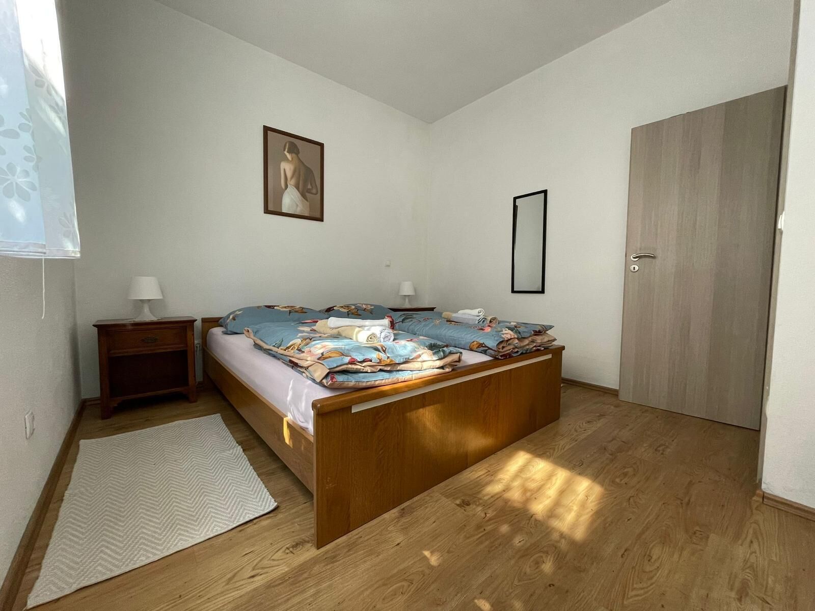 A bedroom with a wooden bed, a nightstand, a bedside table, a mirror, and a door.