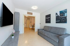 A modern living room with a grey sofa, a flat-screen TV mounted on the wall, and two Prada posters.