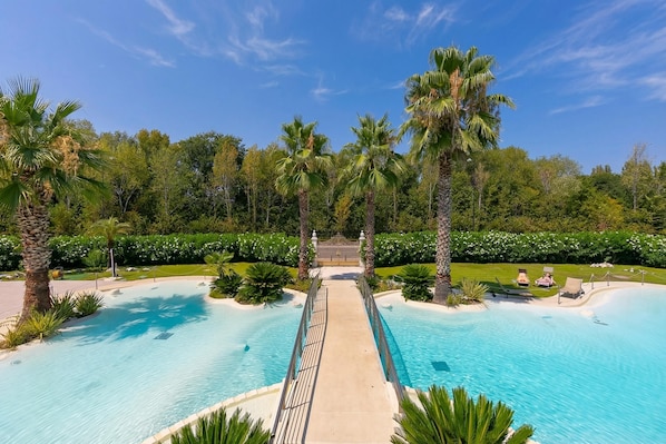 A large outdoor swimming pool with a central walkway, surrounded by palm trees and greenery. - Rivoltella Apt With Pool 600m From Lake (Desenzano del Garda)