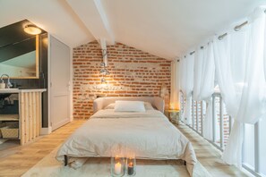 Een slaapkamer met een bakstenen muur, een bed, een nachtkastje, een spiegel en een raam met gordijnen.