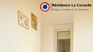 A modern living room with a flat-screen TV, a small table with a lamp, a chair, and a window with blinds. - Résidence La Cocarde (Bourges)