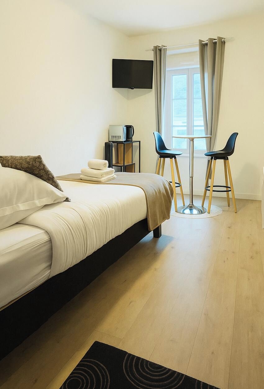 A hotel room with a bed, a TV mounted on the wall, a small table with two chairs, and a window with curtains.