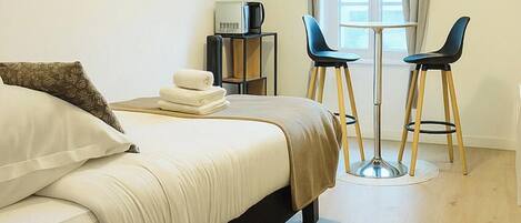 A hotel room with a bed, a TV mounted on the wall, a small table with two chairs, and a window with curtains.