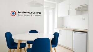 A modern kitchen with white cabinets, a built-in oven, and a dishwasher. There is a round table with blue chairs in the foreground. - Résidence La Cocarde (Bourges)