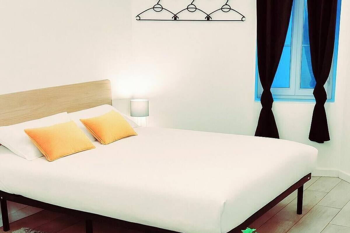 A neatly made bed with white and yellow pillows, a bedside table with a lamp, a black plant on a brown table, and a window with black curtains.