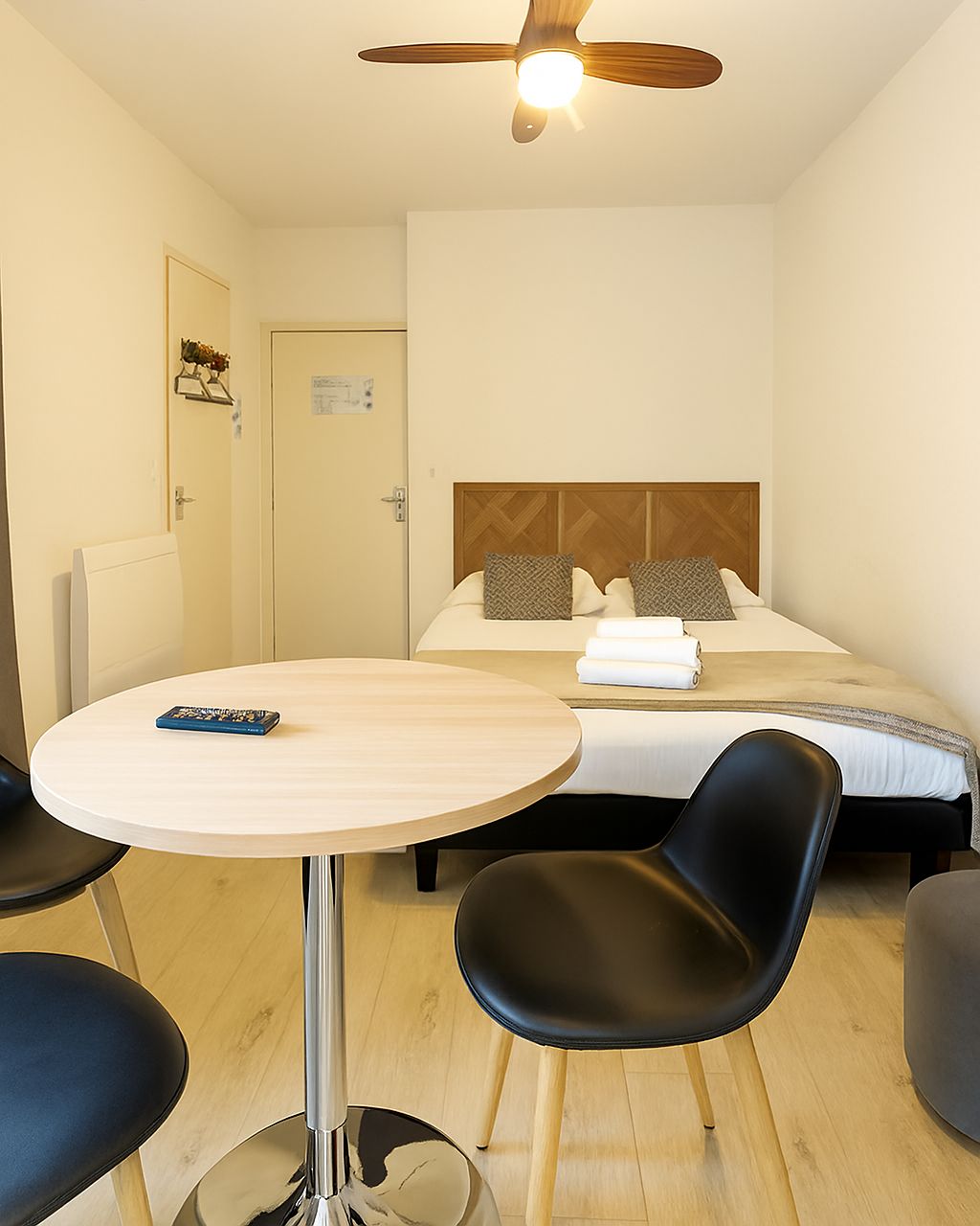 Habitación de hotel con cama, una mesa redonda, dos sillas, un ventilador de techo y una puerta con perchero.