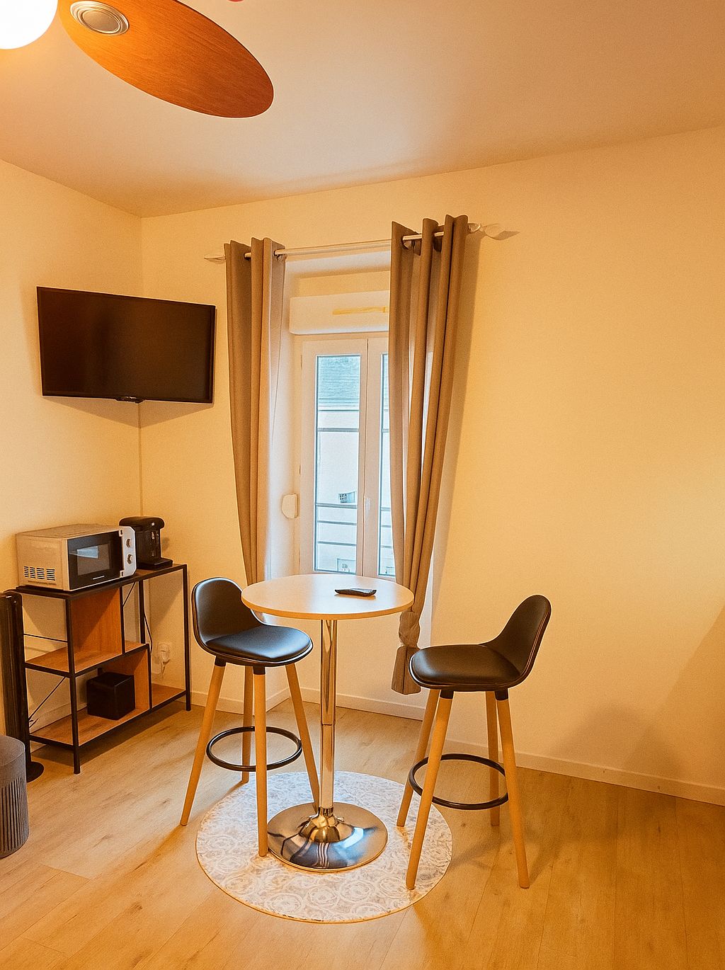 A compact living space with a dining area, a flat-screen TV mounted on the wall, and a window with curtains.