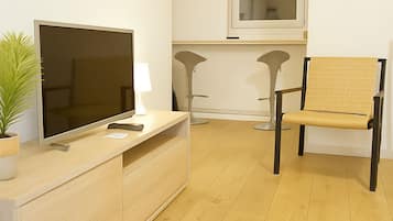 A modern living room with a flat-screen TV, a small table with a lamp, a chair, and a window with blinds.