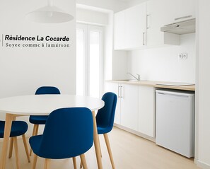 A modern kitchen with white cabinets, a built-in oven, and a dishwasher. There is a round table with blue chairs in the foreground. - Résidence La Cocarde (Bourges)
