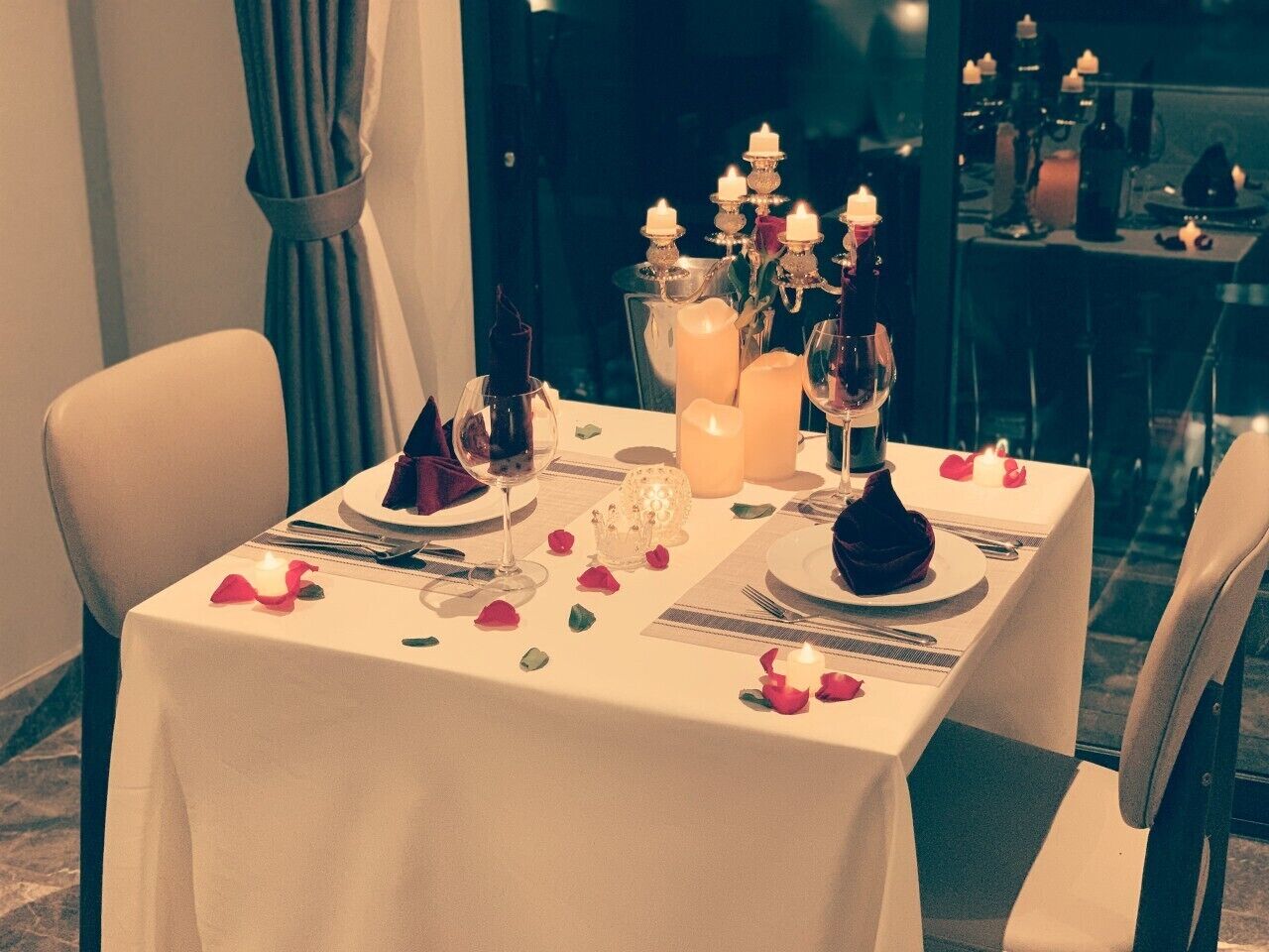 Una mesa de comedor decorada con velas, copas y pétalos de rosa, evocando una cena romántica o íntima.