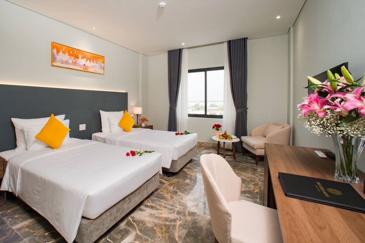 Hotelkamer met twee bedden, een bureau, een stoel, een klein tafeltje met bloemen, een schilderij aan de muur en een raam met gordijnen.