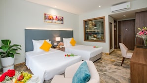 A hotel room with two beds, a desk, a chair, a potted plant, and a painting on the wall. - Phu Thang Grand Hotel (Duc Hoa)