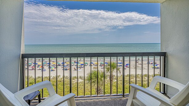 A balcony with two white chairs, a view of a beach with umbrellas, and the ocean. - Turtle Cove, Spectacular Ocean Front 1 bedroom! (Myrtle Beach)