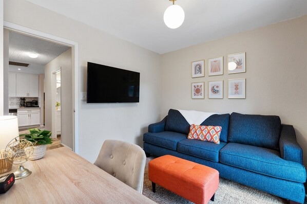Una sala de estar con un sofá azul, un reposapiés naranja, un sillón beige y un televisor de pantalla plana en la pared.