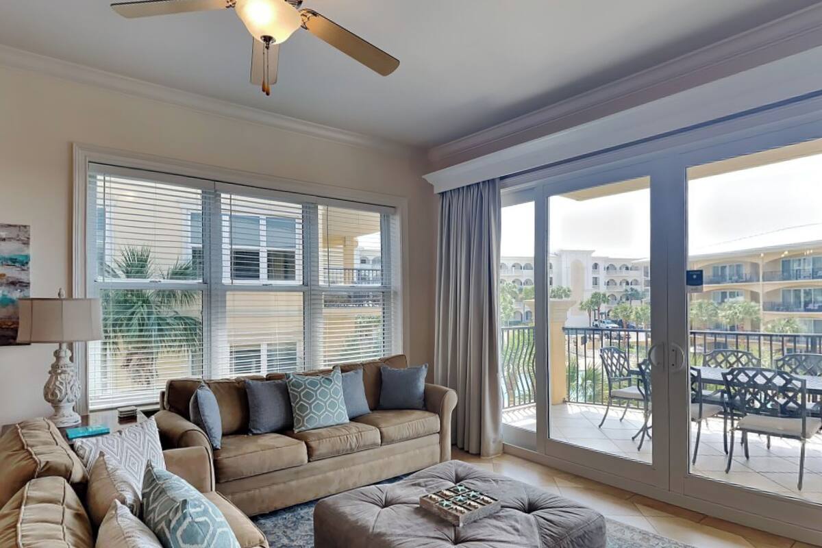 A spacious living room with a large window, a ceiling fan, and a view of a building and palm trees outside.