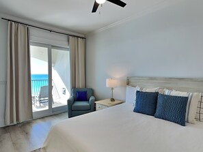 A hotel room with a large bed, a nightstand with a lamp, a chair, and a view of the ocean through the sliding glass door.