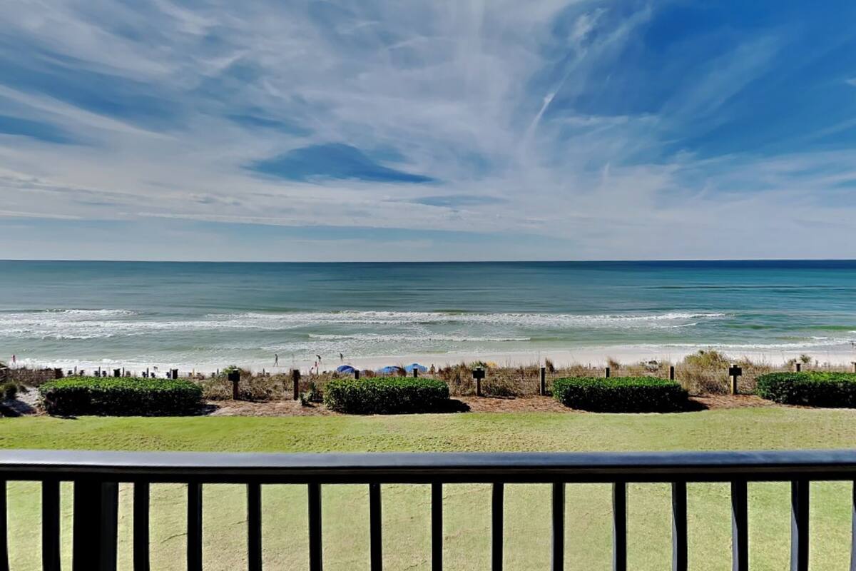 A beach view with a clear sky, ocean waves, and a grassy area with bushes and a fence in the foreground.