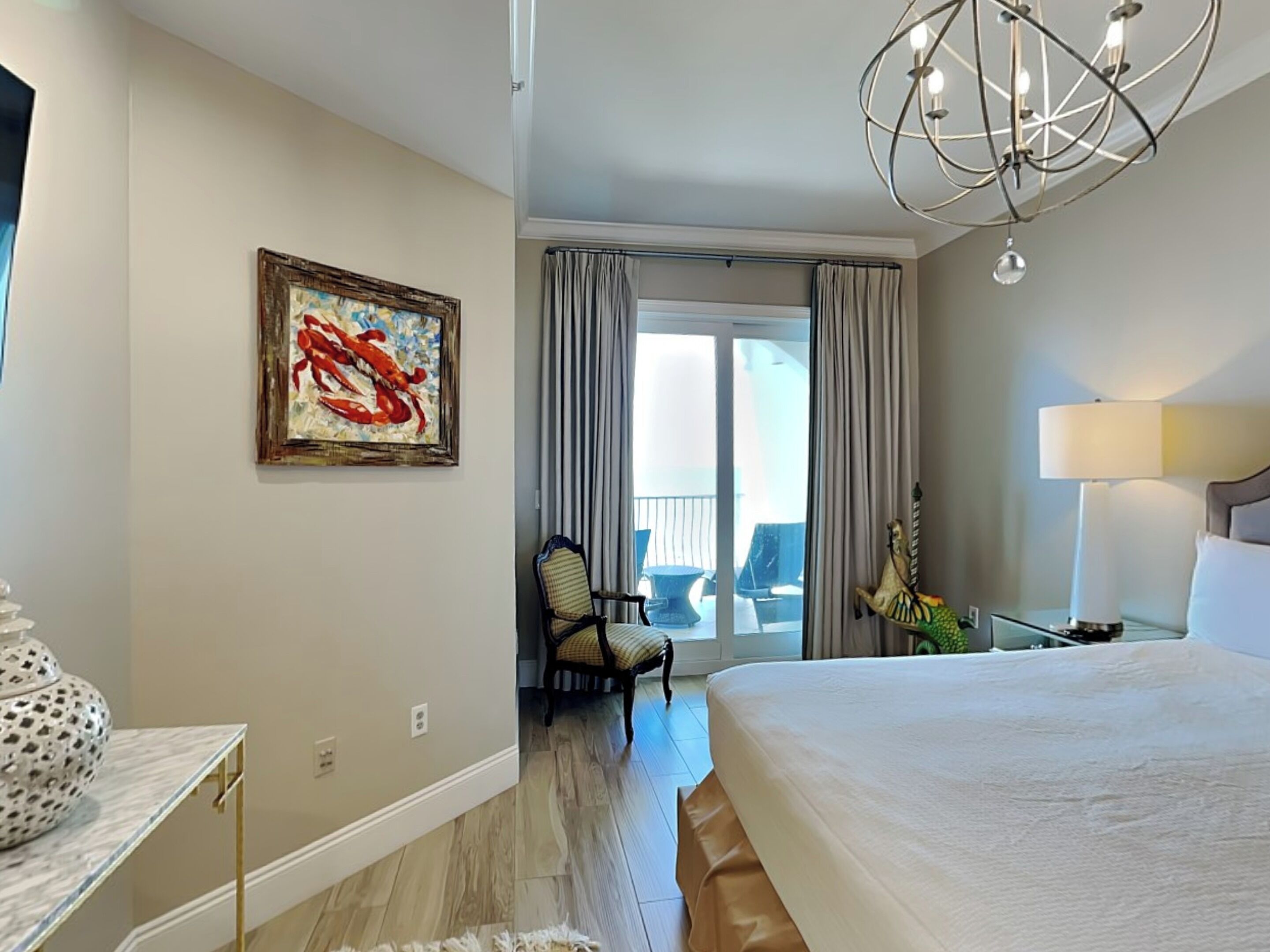 A hotel room with a large bed, a chandelier, a painting, a chair, and a balcony with curtains.