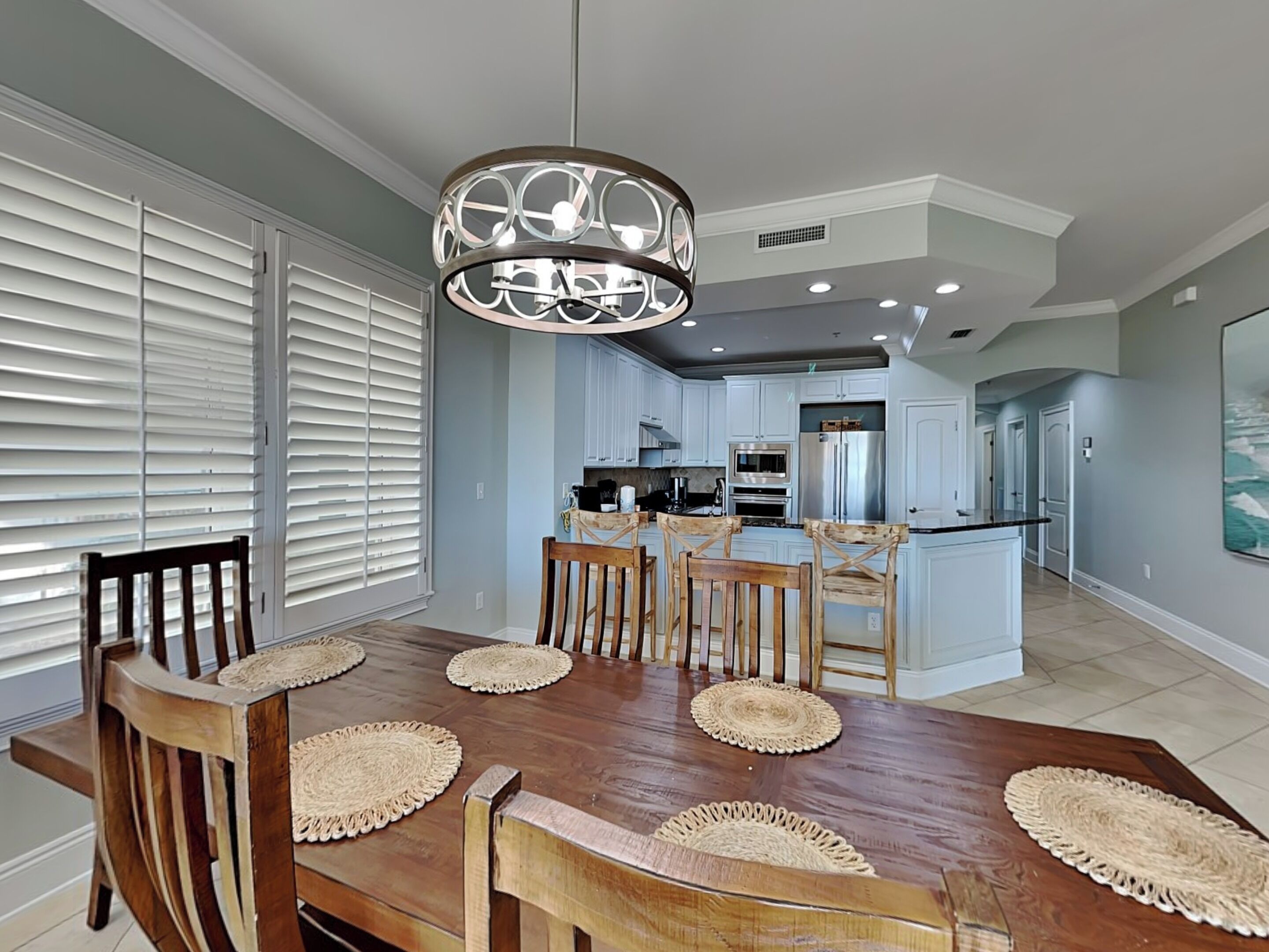 A dining area with a wooden table and chairs, a chandelier, and a kitchen in the background.