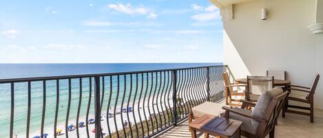 A balcony with a view of the beach and ocean. There are wooden chairs and a small table.