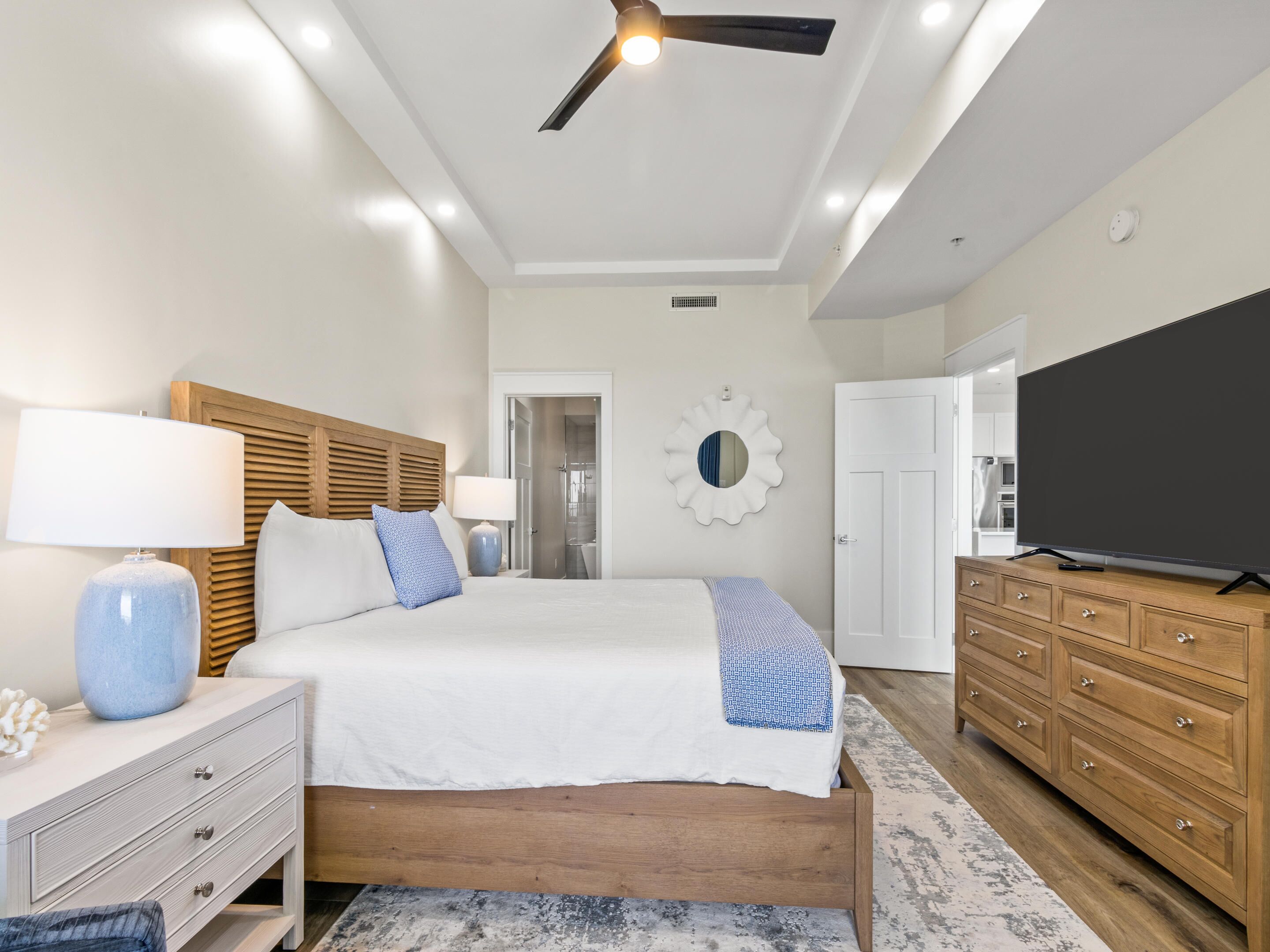 A modern bedroom with a bed, dresser, TV, ceiling fan, and a bathroom door.