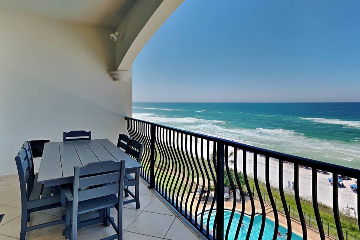 A balcony with a table and chairs, overlooking a pool and the ocean.