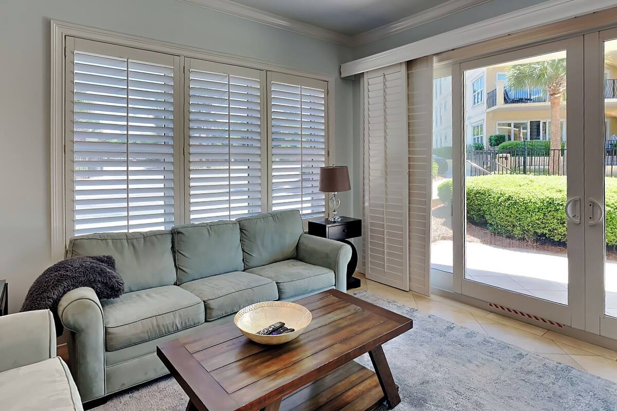 A spacious living room with a light green sofa, a wooden coffee table with a bowl, and a lamp on a side table. Large windows with blinds allow natural light, and a sliding glass door leads to an outdoor area.