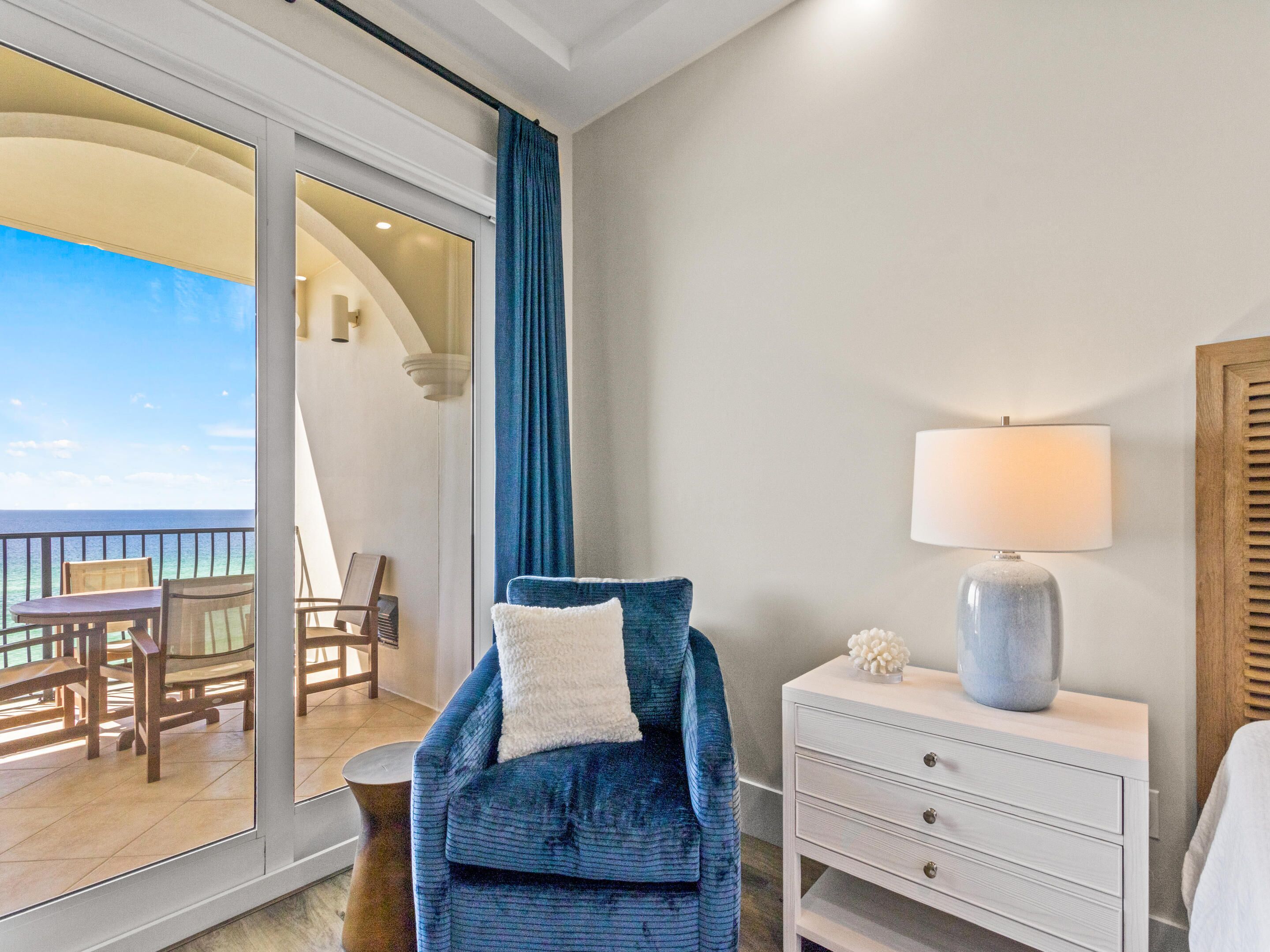 Een kamer met een blauwe fauteuil, een witte nachtkastje met een lamp en uitzicht op de oceaan door een glazen deur.
