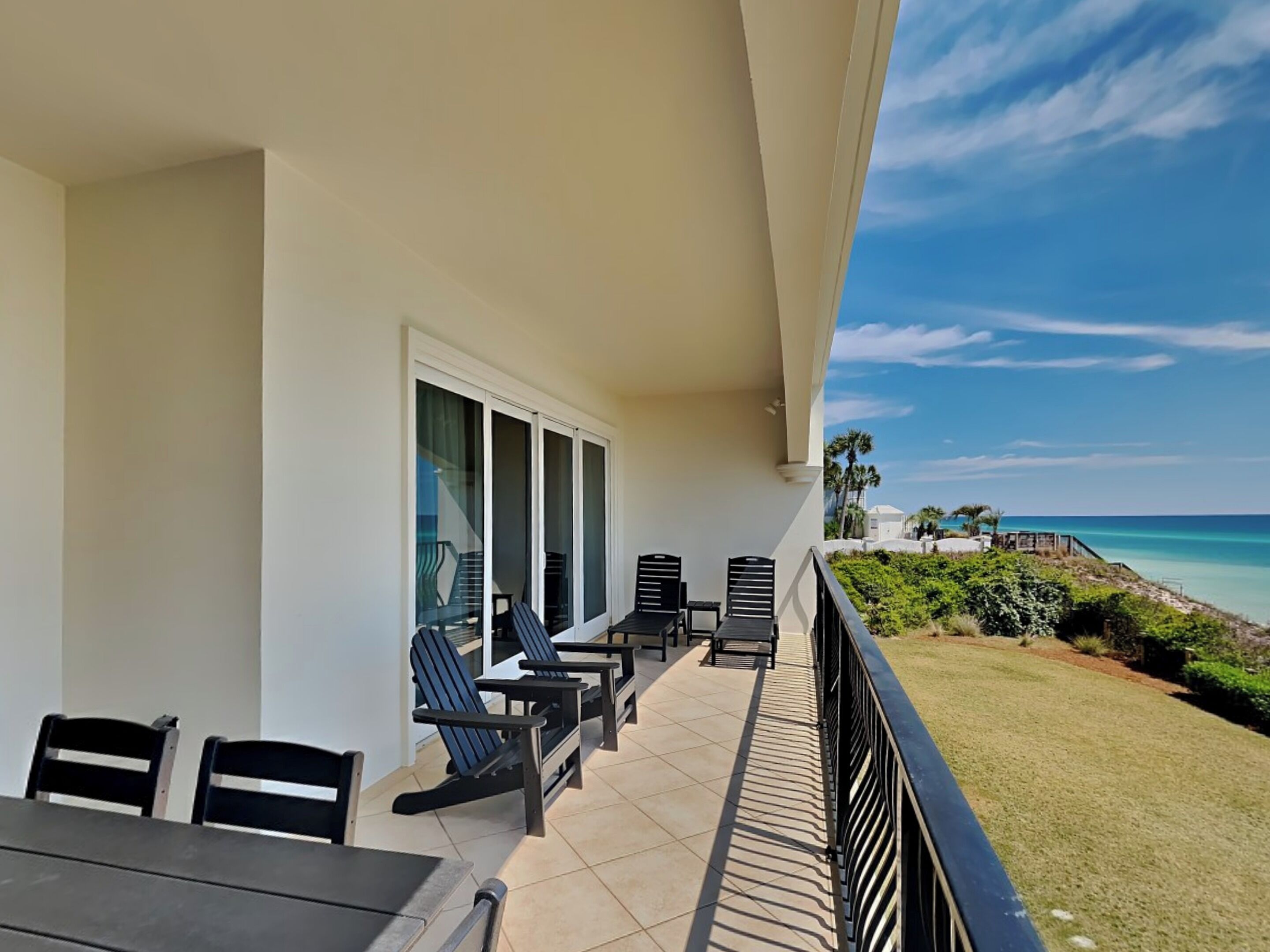 A balcony with a table and chairs, a view of the ocean, and a grassy area with plants.