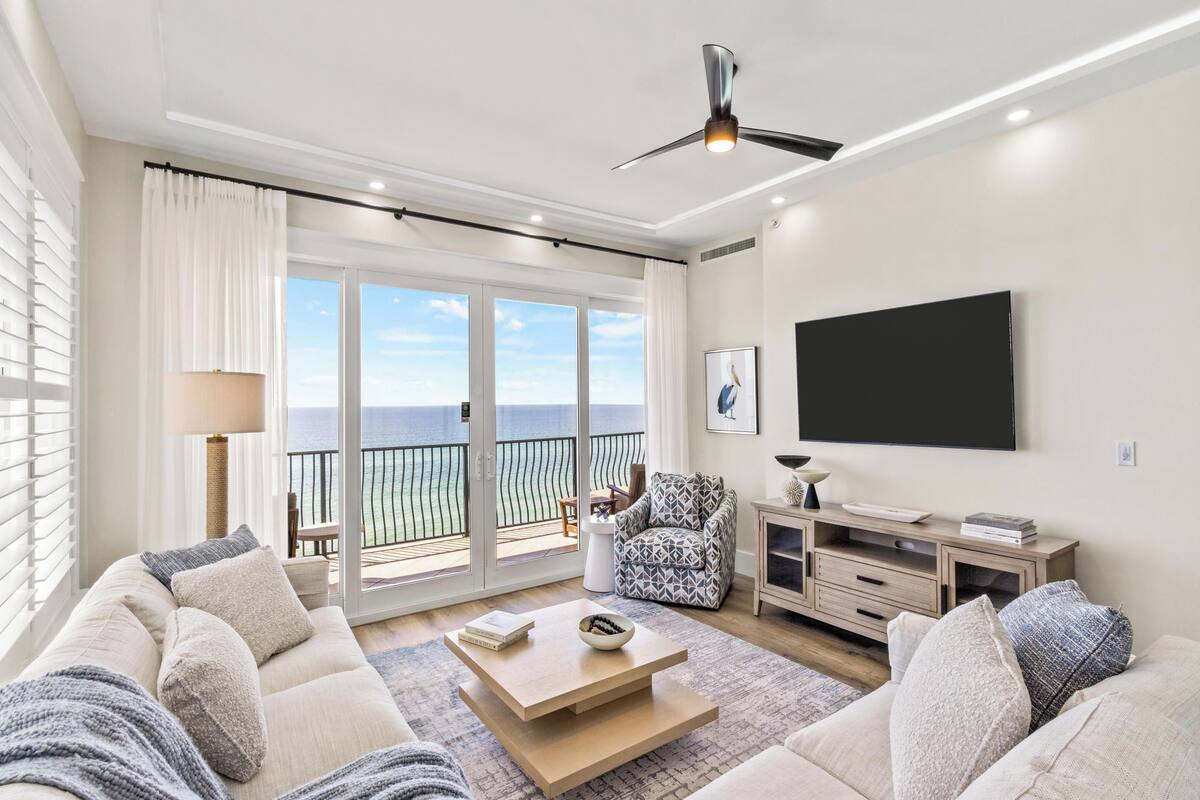 A spacious living room with a balcony, a ceiling fan, a flat-screen TV, and a large window with a view of the ocean.