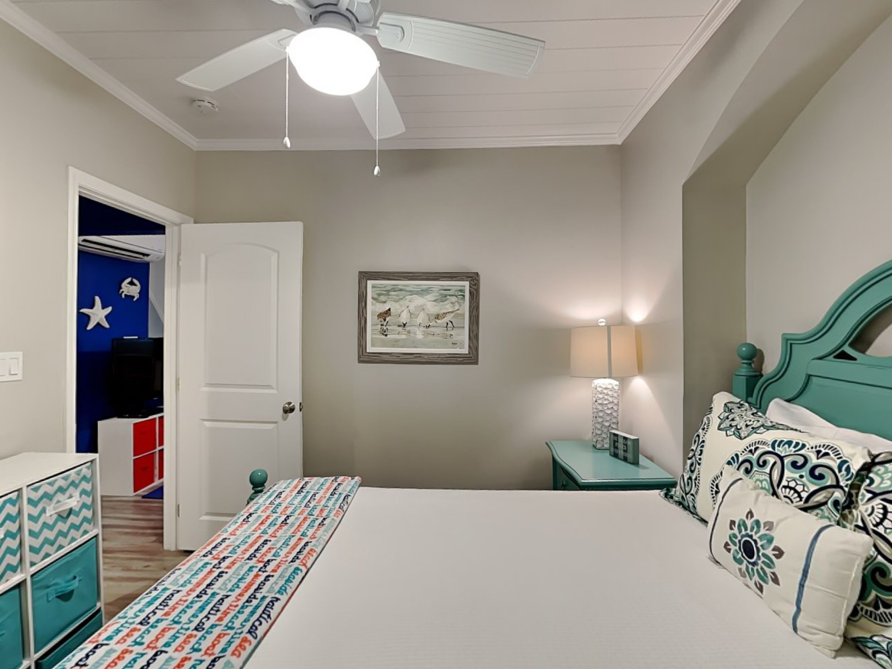 Una camera da letto con un letto, una ventola a soffitto, una lampada da comodino, un quadro appeso al muro e una porta che dà su un'altra stanza.