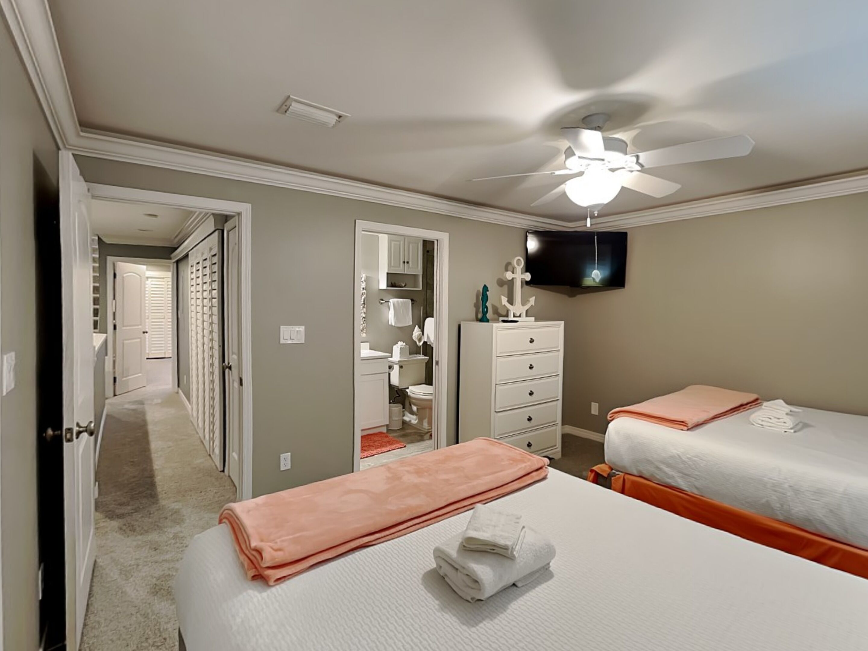 A hotel room with two beds, a ceiling fan, a TV, a bathroom with a mirror, and a hallway leading to another room.