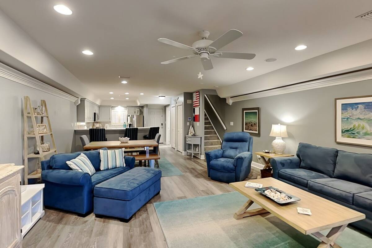 A spacious living room with a blue sofa, a matching ottoman, a wooden coffee table, and a blue armchair. A staircase leads to a kitchen area in the background.