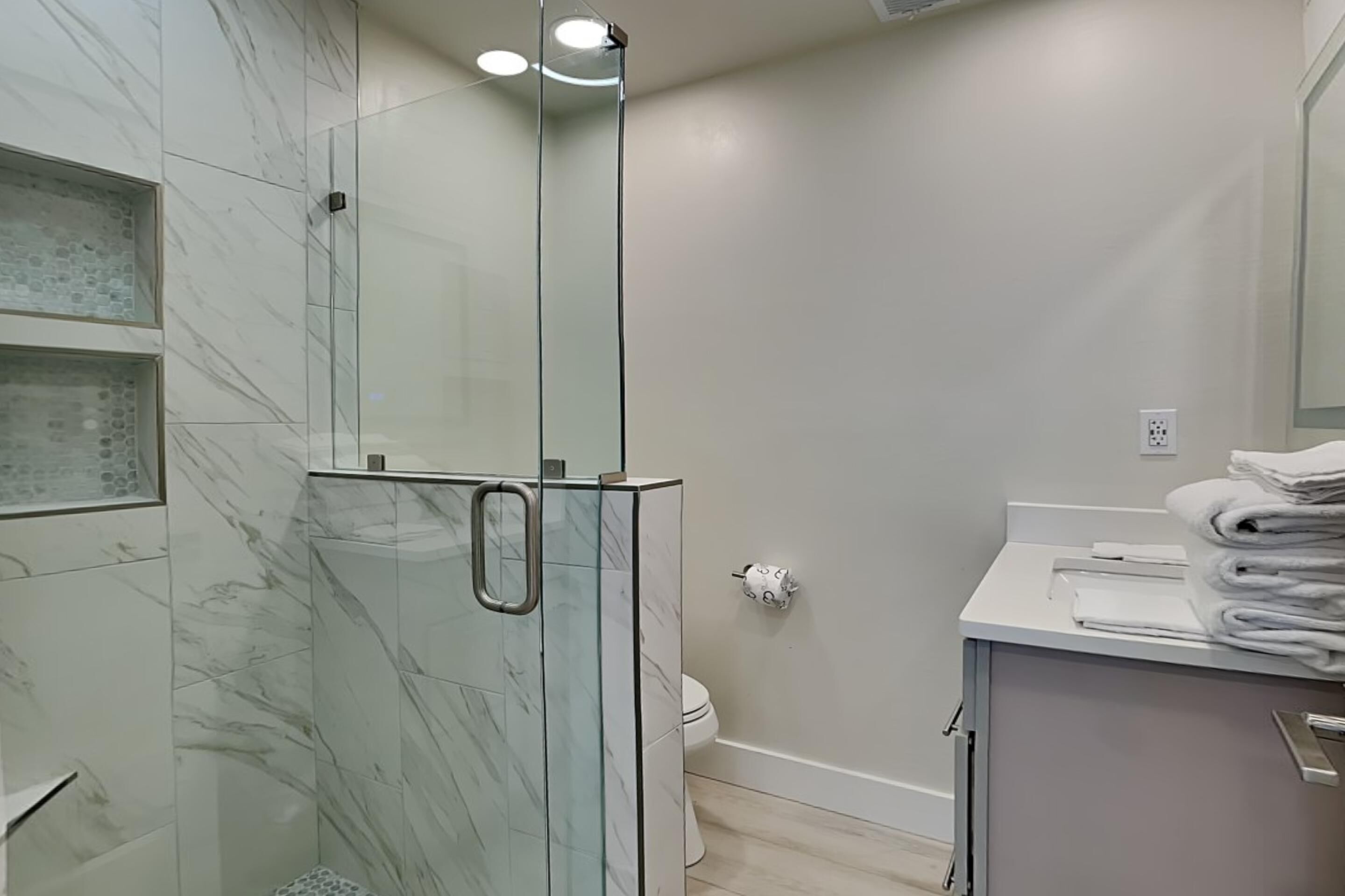 Une salle de bain moderne dotée d’une douche à parois de verre, d’un mur recouvert de carreaux de marbre, de toilettes et d’un coin lavabo avec des piles de serviettes.