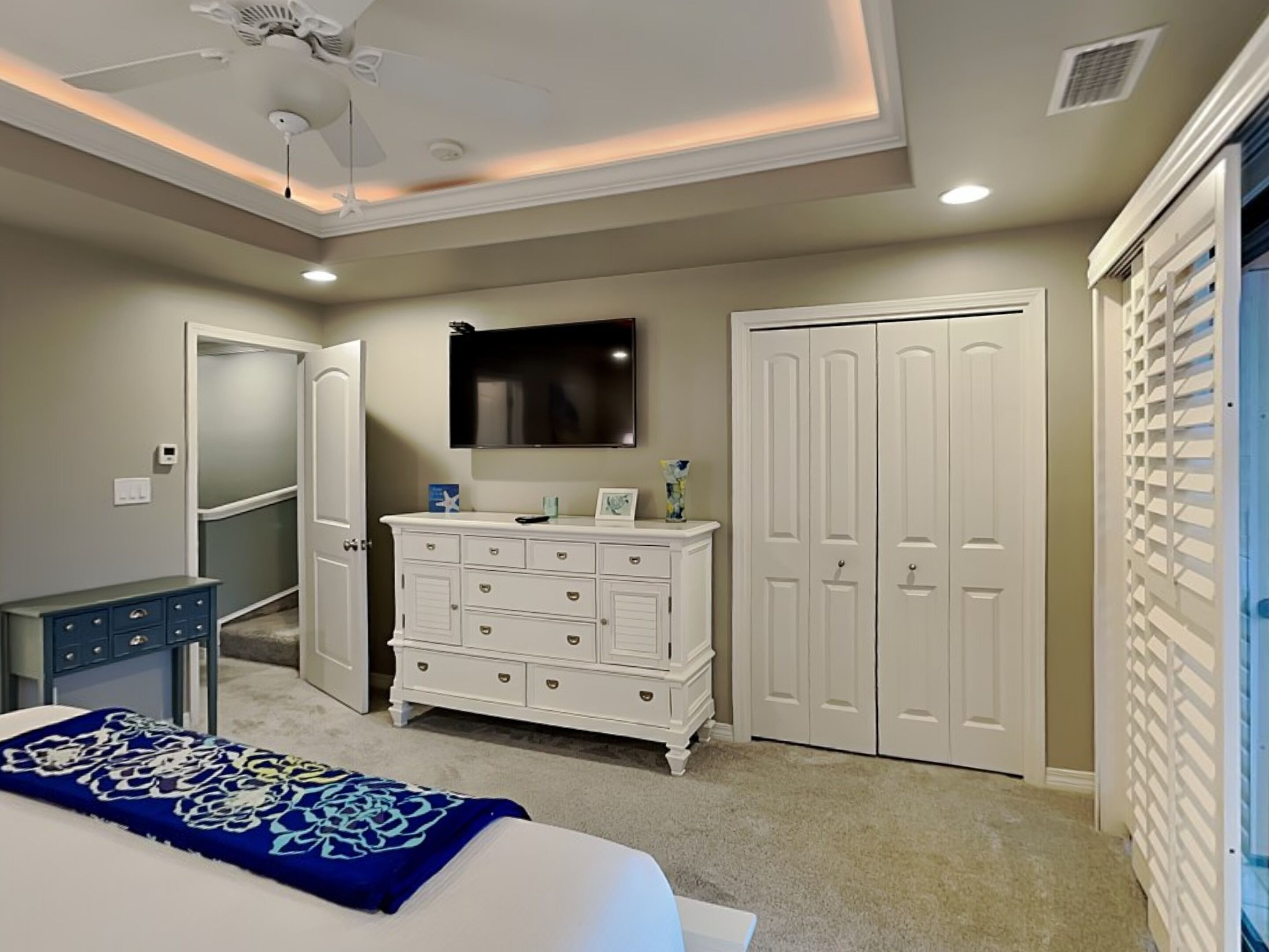A bedroom with a bed, dresser, wardrobe, ceiling fan, and a mounted TV.